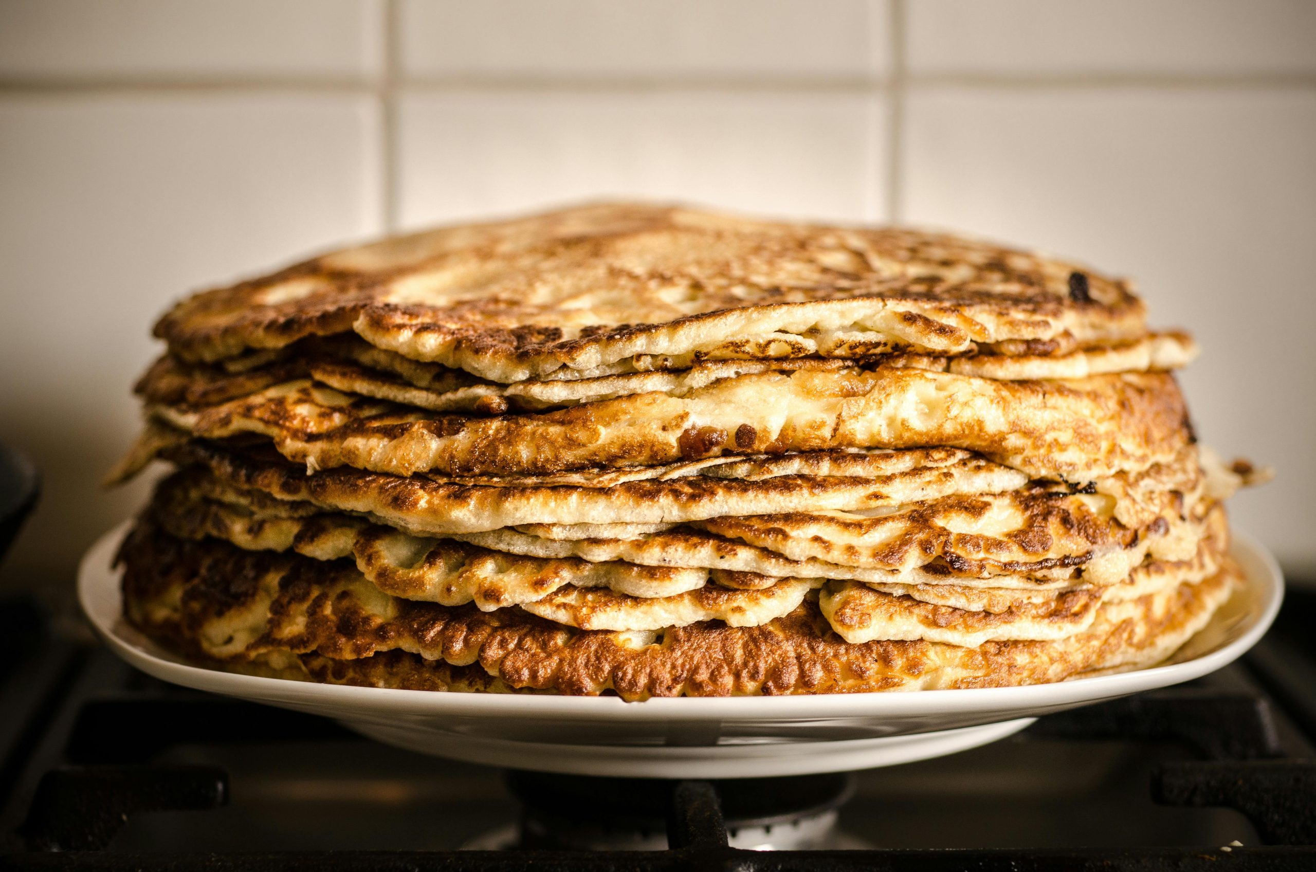 Ett fat med en hög stapel nystekta tunna pannkakor, gyllenbruna och frasiga, stående på ett bord i ett kök.